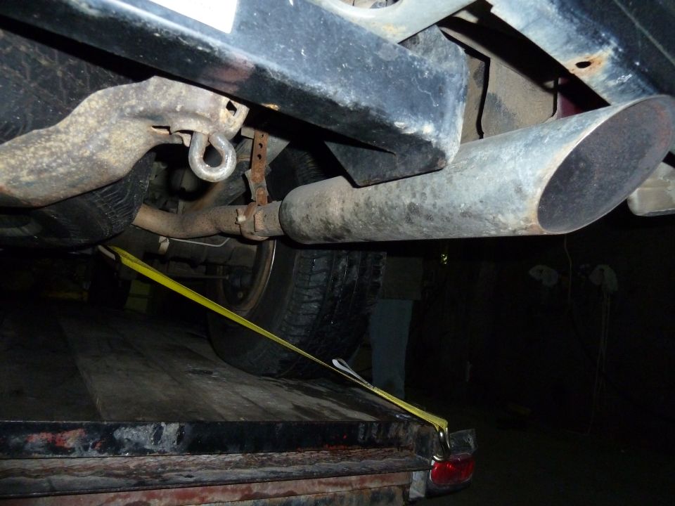 Underside of a vehicle showing exhaust pipe, suspension components, and a yellow tie-down strap