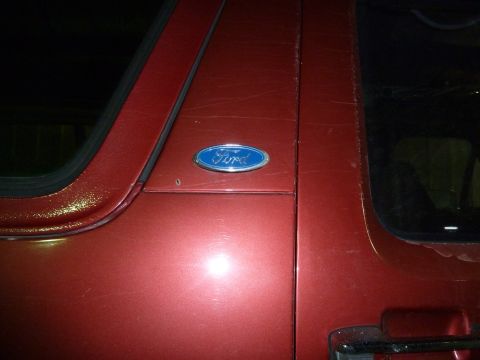 1993 Ford C-150 (Centurion Conversions) - Close-up of a red vehicle's side door, showing the Ford emblem and surface imperfections.