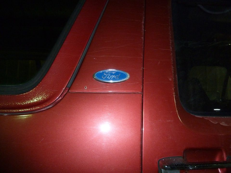 Red vehicle door with a blue Ford oval emblem. Scratches are visible on the paint