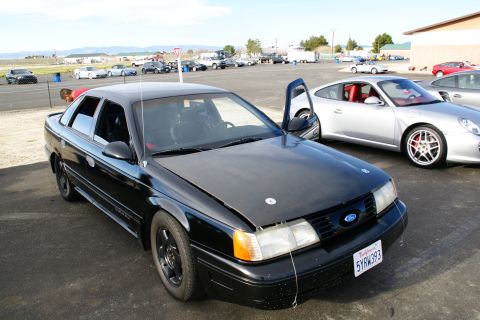 1995 Ford Taurus - A black sedan with racing modifications is parked on asphalt, with a silver sports car to its right.