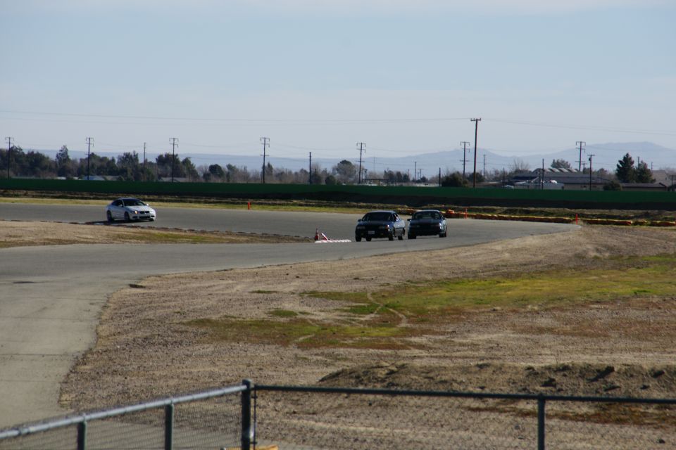 Three cars are on a racetrack, two dark sedans are close together on a curve, and a white sedan is further back on the track