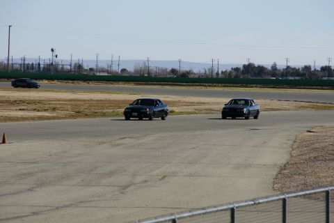 1995 Ford Taurus - Two cars are on a race track under a clear sky with power lines visible in the distance.