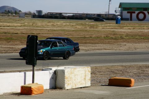 1995 Ford Taurus - A teal sedan and a dark sedan race on a track with a building in the background.