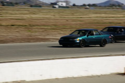 1995 Ford Taurus - A teal 1995 Ford Taurus with a black hood races on a track, followed by another vehicle.