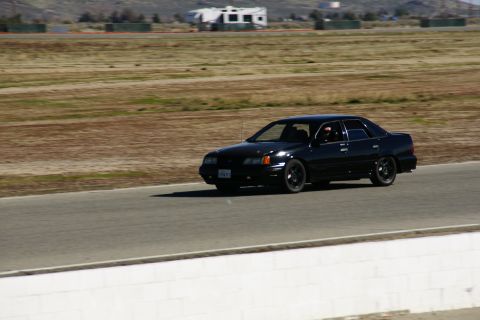 1995 Ford Taurus - A black sedan drives on a race track with dry grass and buildings in the background.
