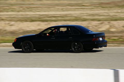 1995 Ford Taurus - A black 1995 Ford Taurus sedan is captured in motion on a race track, viewed from the side.
