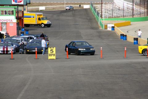1995 Ford Taurus - A dark sedan drives on a track, passing a 5 MPH speed limit sign. Other cars and a truck are visible in the background.