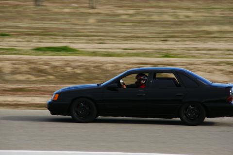 1995 Ford Taurus - A black sedan drives on a track with a driver in a helmet visible through the window.