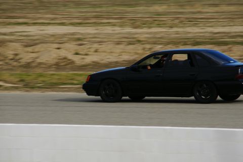 1995 Ford Taurus - A black 1995 Ford Taurus sedan is captured in profile as it drives on a racetrack.