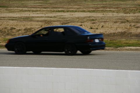 1995 Ford Taurus - 1995 Ford Taurus sedan captured in motion on a paved track with dry grass and dirt in the background.