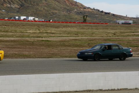 1995 Ford Taurus - A dark green sedan, likely a 1995 Ford Taurus, drives on a racetrack with a yellow vehicle partially visible to the left.