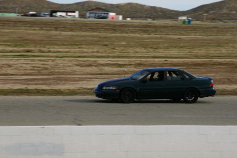 1995 Ford Taurus - A dark green 1995 Ford Taurus sedan is captured in profile while driving on a racetrack.