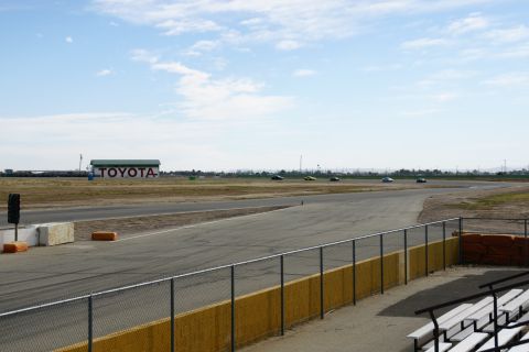 1995 Ford Taurus - Several cars are racing on a track with a large Toyota building visible in the distance under a partly cloudy sky.