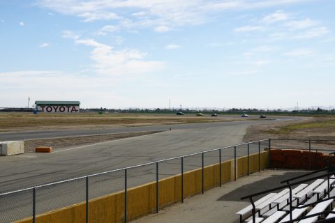 1995 Ford Taurus - Several cars are on a race track under a partly cloudy sky, with a large Toyota building visible in the distance.