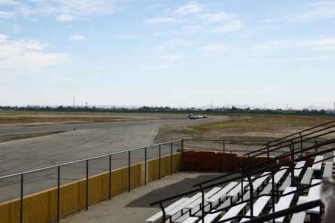 1995 Ford Taurus - Two cars drive on a racetrack under a partly cloudy sky, with empty grandstands visible.