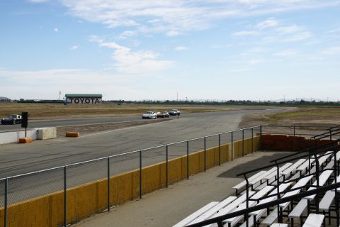 1995 Ford Taurus - Multiple cars race on a track with a large Toyota building visible in the distance under a partly cloudy sky.