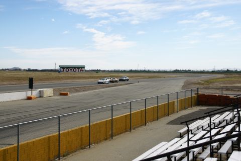 1995 Ford Taurus - Three cars race on a track with a Toyota building visible in the distance under a partly cloudy sky.
