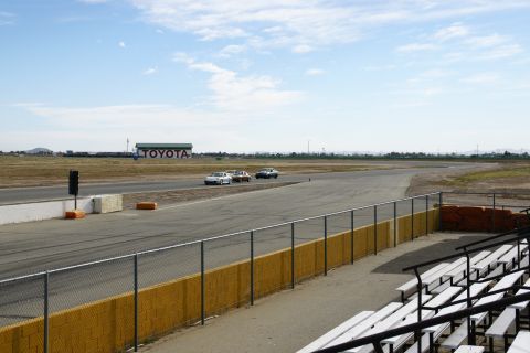 1995 Ford Taurus - Cars race on a track with a large Toyota building visible in the distance under a cloudy sky.