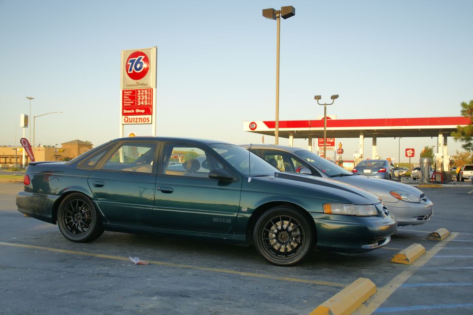 Green 1995 Ford Taurus with aftermarket wheels parked next to another car at a gas station