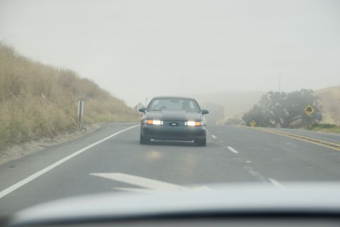 1995 Ford Taurus - A dark sedan drives down a highway shrouded in fog, its headlights illuminated.
