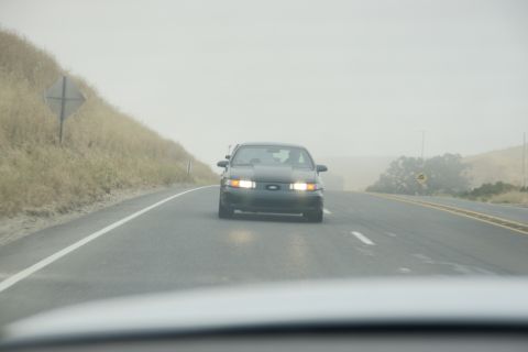 1995 Ford Taurus - A dark sedan drives down a highway on a hazy day, its headlights illuminated.