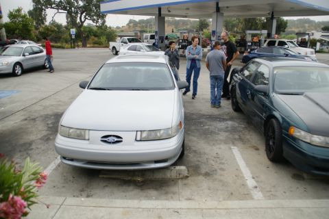 1995 Ford Taurus - A silver sedan is parked at a gas station with people standing nearby.