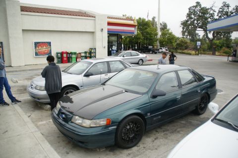 1995 Ford Taurus - A dark green sedan with a black hood is parked next to a silver sedan at a gas station.