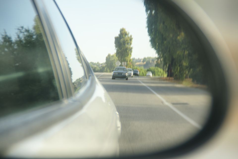 View in side mirror shows cars driving on a road with trees and hills