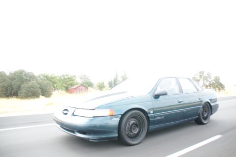 1995 Ford Taurus - A green 1995 Ford Taurus sedan is captured in motion on a highway, with a rural landscape visible to the side.