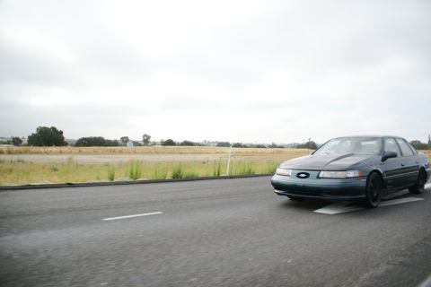1995 Ford Taurus - A dark green sedan is captured from the front 3/4 angle while driving on a highway.