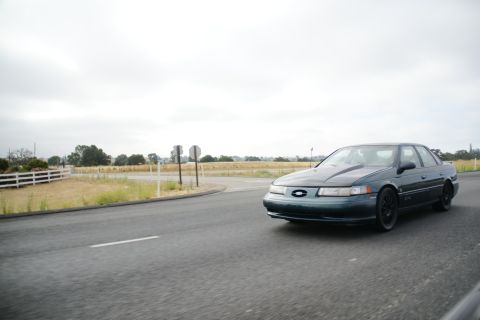1995 Ford Taurus - A dark green 1995 Ford Taurus sedan is captured in a 3/4 front view as it drives along a paved road under an overcast sky.
