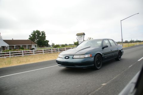 1995 Ford Taurus - A dark green sedan is captured in motion on a highway, with a white fence and buildings visible to the left.