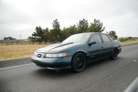 1995 Ford Taurus - A 1995 Ford Taurus sedan is captured in motion on a highway under an overcast sky.
