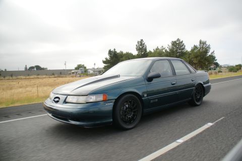 1995 Ford Taurus - A dark green 1995 Ford Taurus sedan is captured in motion on a highway under an overcast sky.