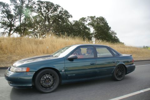 1995 Ford Taurus - A green sedan with aftermarket wheels drives down a highway flanked by dry grass and trees.