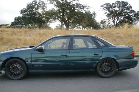 1995 Ford Taurus - A green 1995 Ford Taurus sedan drives down a road with dry grass and trees in the background.
