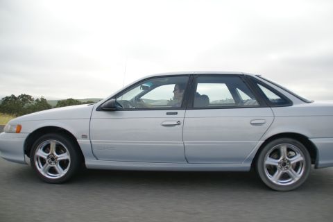 1995 Ford Taurus - A silver 1995 Ford Taurus sedan is captured in a side profile shot while driving on a road.