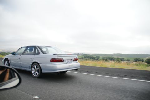 1995 Ford Taurus - A silver sedan drives down a highway with a scenic rural landscape under a cloudy sky.