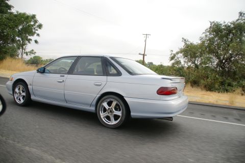 1995 Ford Taurus - Silver sedan with aftermarket modifications drives down a highway on an overcast day.