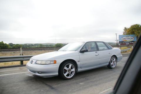 1995 Ford Taurus - A silver 1995 Ford Taurus sedan is seen driving on a highway, captured from a side angle.