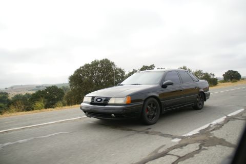 1995 Ford Taurus - 1995 Ford Taurus sedan captured in motion on a highway with overcast skies.