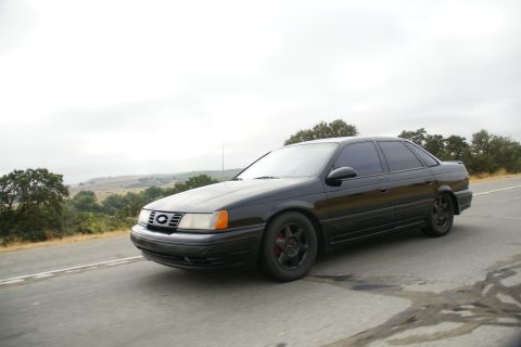 1995 Ford Taurus - A black sedan is captured in motion on a highway, with a scenic landscape visible behind it.