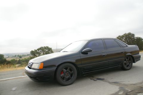 1995 Ford Taurus - A black 1995 Ford Taurus sedan is seen from a 3q front angle on a road with a scenic background.