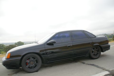 1995 Ford Taurus - A black sedan is shown from the side, driving on a road with a hazy sky and distant hills.