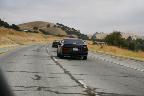 1995 Ford Taurus - Two sedans drive along a cracked highway with rolling hills in the background.