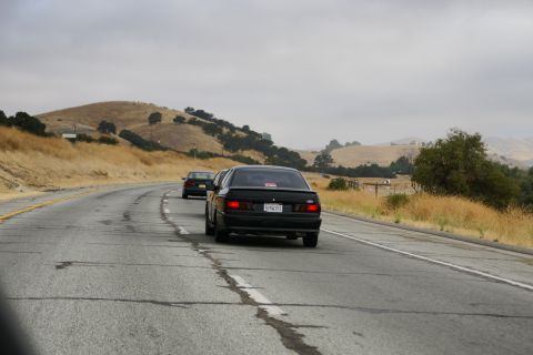 1995 Ford Taurus - Two black sedans travel down a highway through a dry, hilly landscape under an overcast sky.