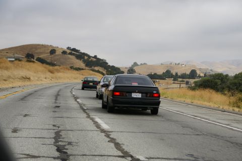 1995 Ford Taurus - A black sedan is seen driving on a cracked highway with other vehicles in a hilly, dry landscape.
