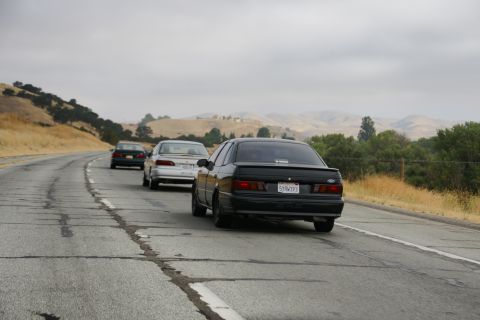 1995 Ford Taurus - Three cars, including a black 1995 Ford Taurus, travel down a highway on a cloudy day.