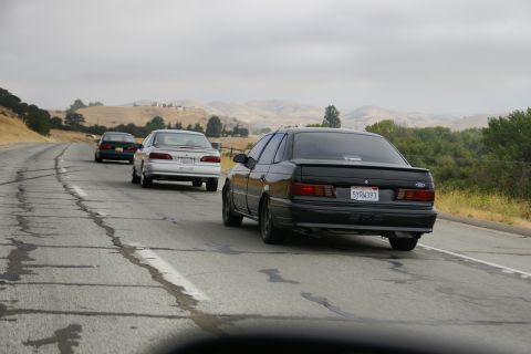 1995 Ford Taurus - A black Ford Taurus drives on a highway behind other vehicles under a cloudy sky.