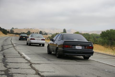 1995 Ford Taurus - Three cars, including a black 1995 Ford Taurus, travel down a cracked highway under a cloudy sky.